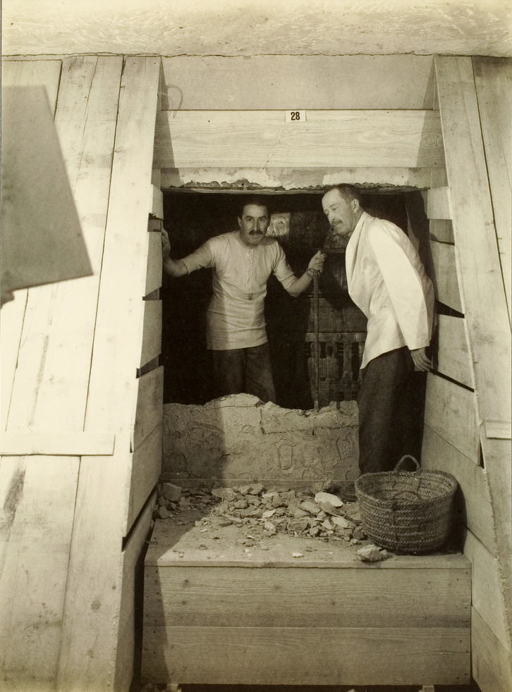 Fotografía de Carter con chaqueta clara y un capataz egipcio junto a un tabique semiderruido en un entarimado de madera