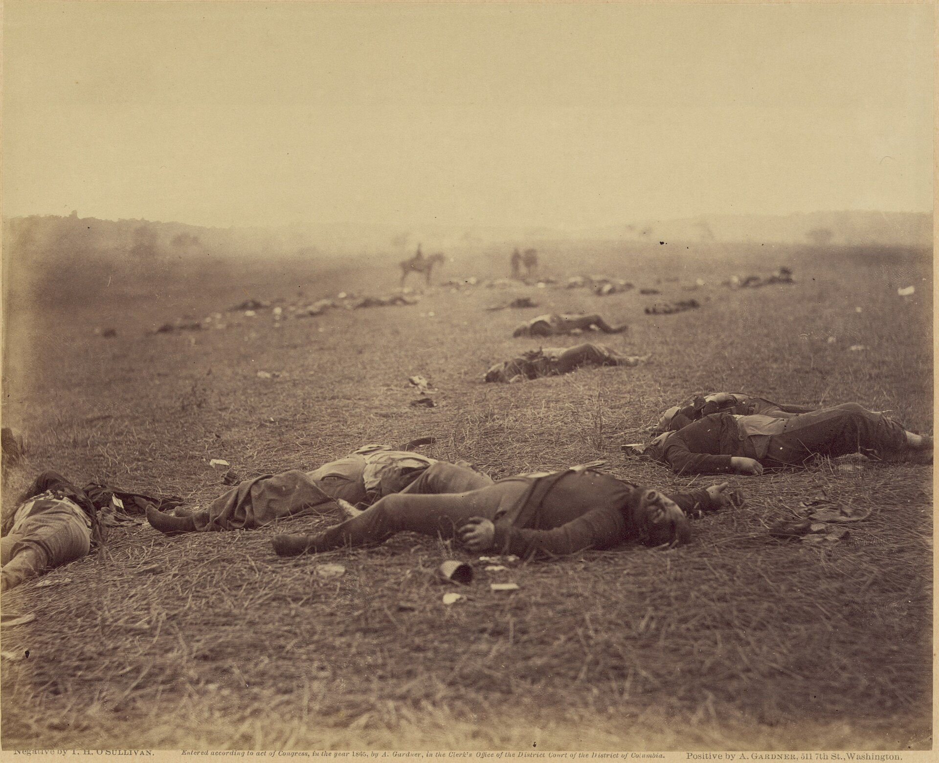 Fotografía de 1863 de un campo con varios cadáveres de soldados tendidos en primer plano y jinetes difuminados al fondo