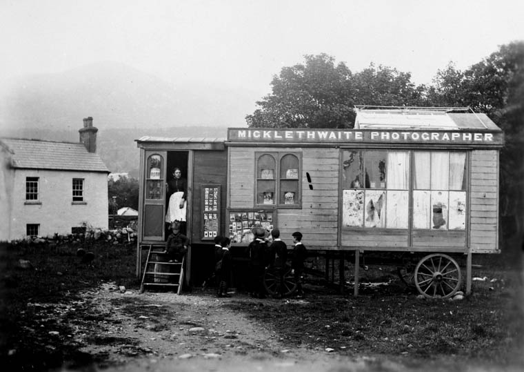 Fotografía del siglo XIX de una caseta de madera con ruedas y el rótulo «Micklethwaite Photographer», con muestras de retratos expuestas y varias personas en la puerta