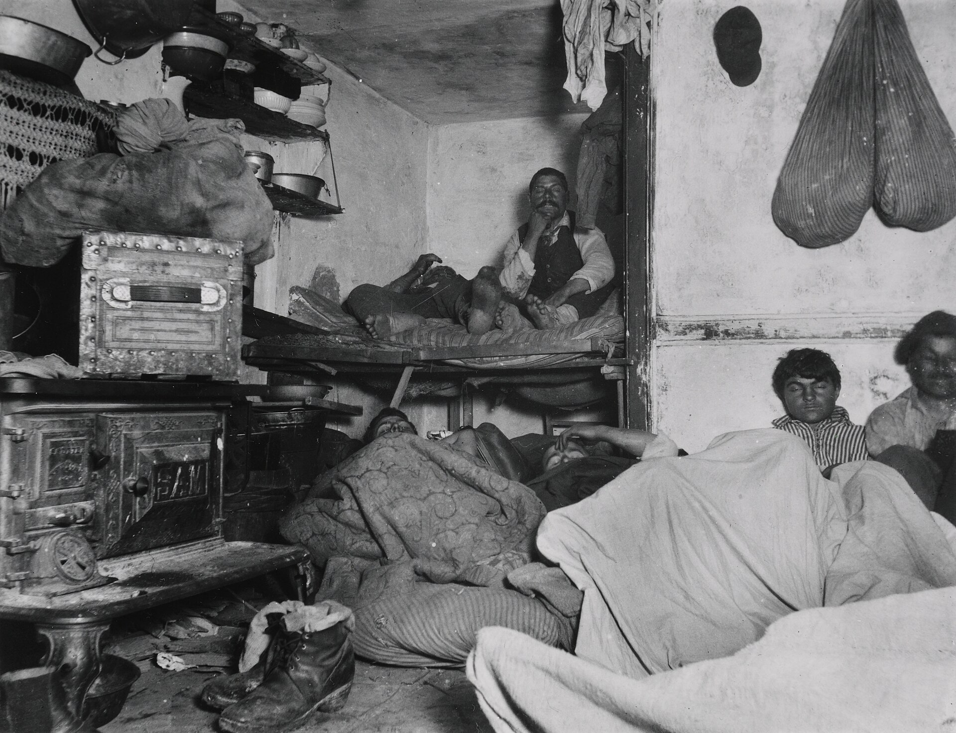 Fotografía nocturna de un sótano con varias personas apiñadas en literas de tablas y ropa colgando de las paredes