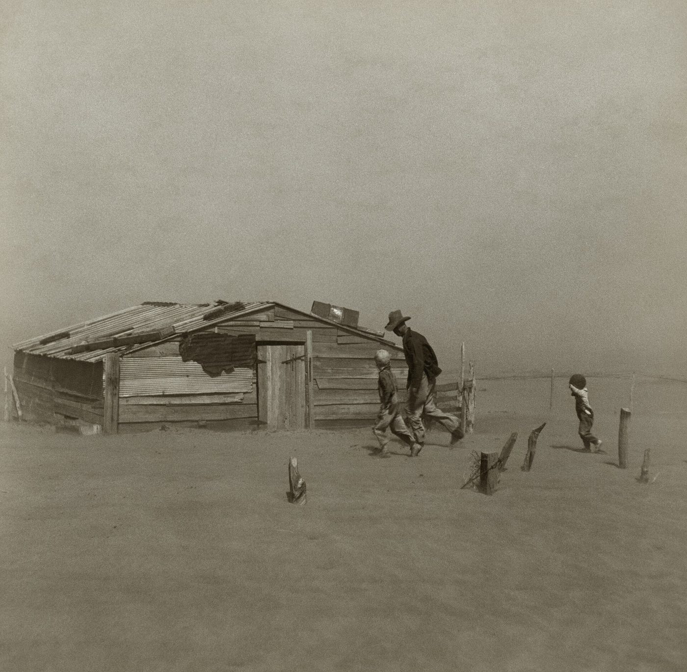 Fotografía de 1936 de un hombre y dos niños caminando encorvados hacia una cabaña medio enterrada mientras el viento levanta el polvo