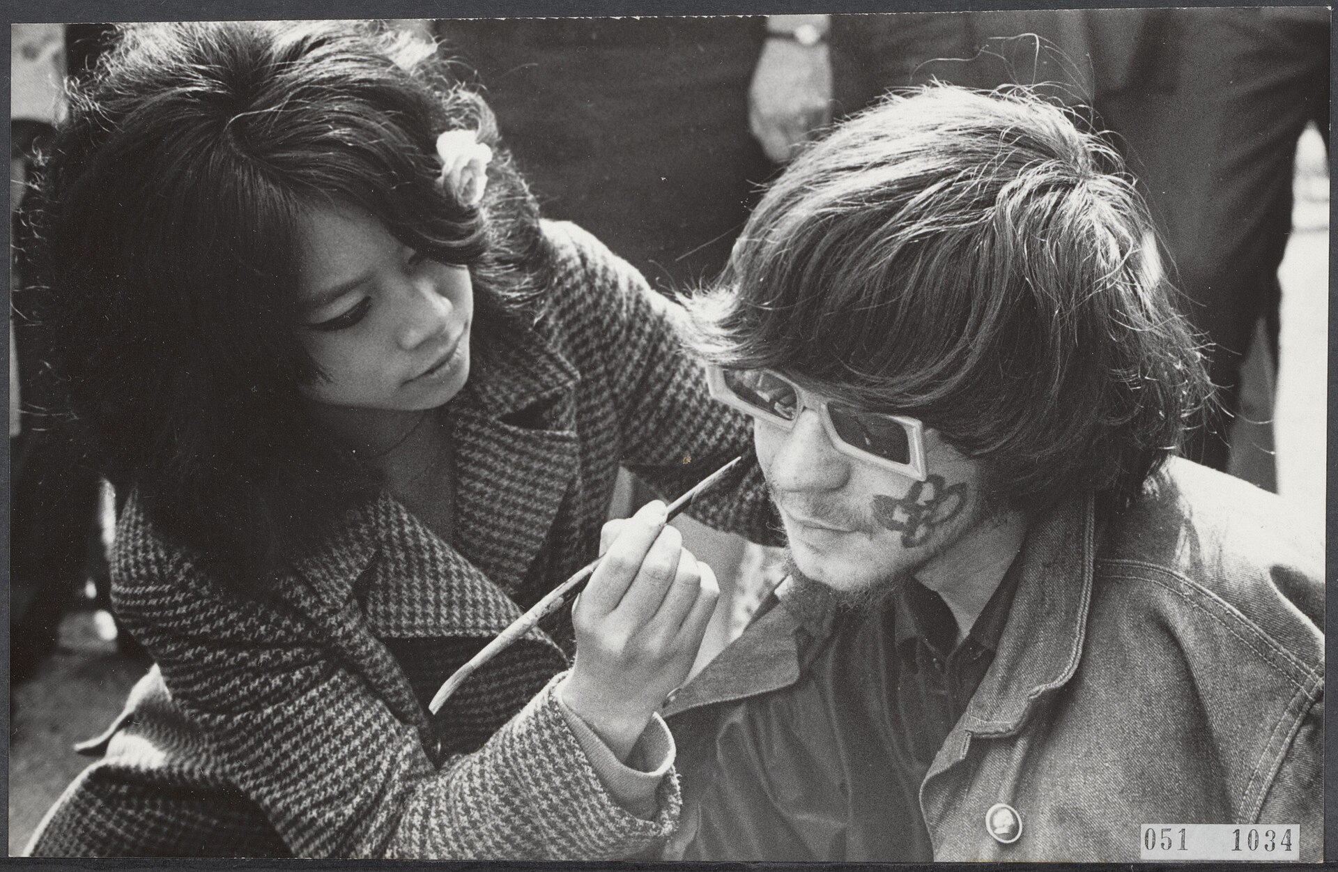 Fotografía en blanco y negro de finales de los años sesenta de una joven pintando con un pincel la cara de un joven de pelo largo