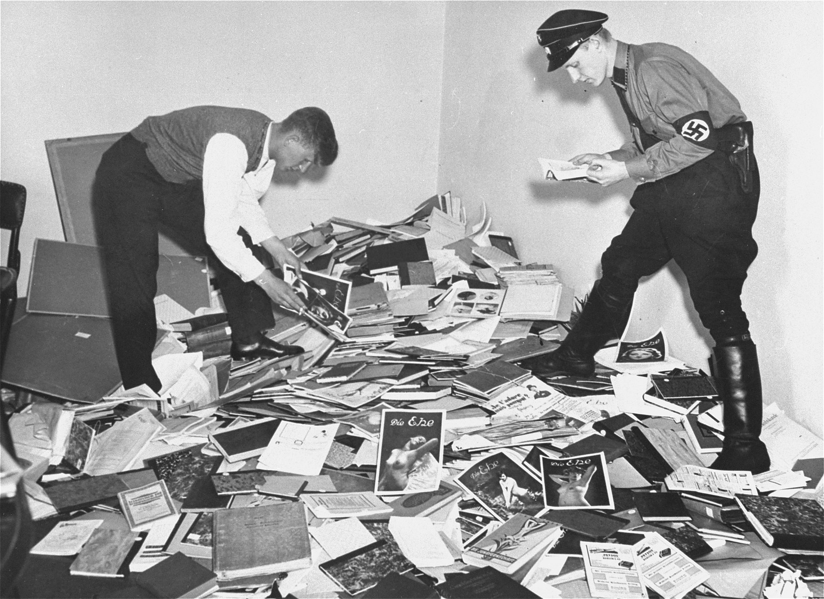 Fotografía de 1933 de dos hombres, uno con brazalete, revolviendo montones de papeles y libros esparcidos por el suelo de una sala