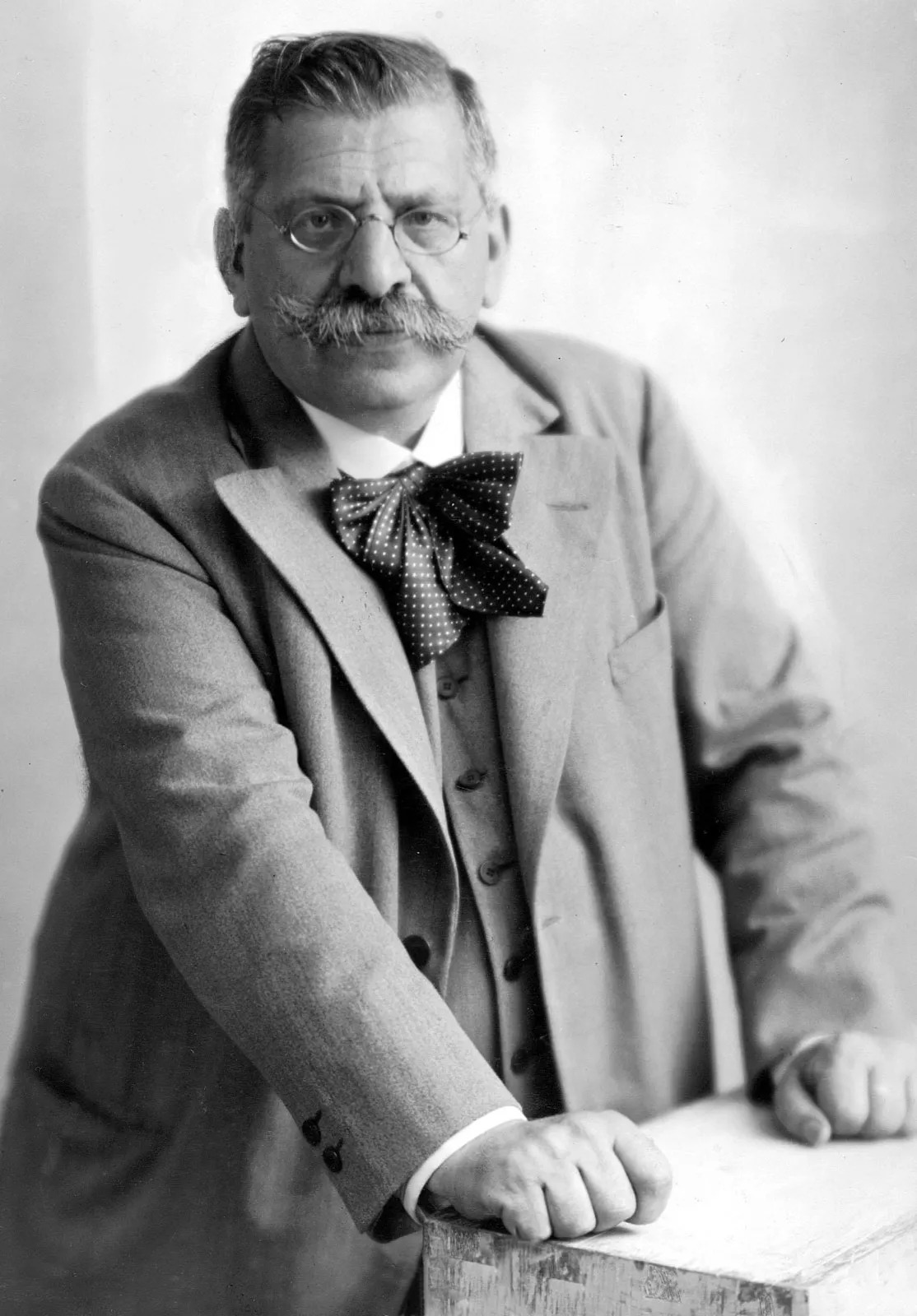 Retrato fotográfico de un hombre de mediana edad con gafas redondas y bigote poblado, sentado ante una mesa de despacho