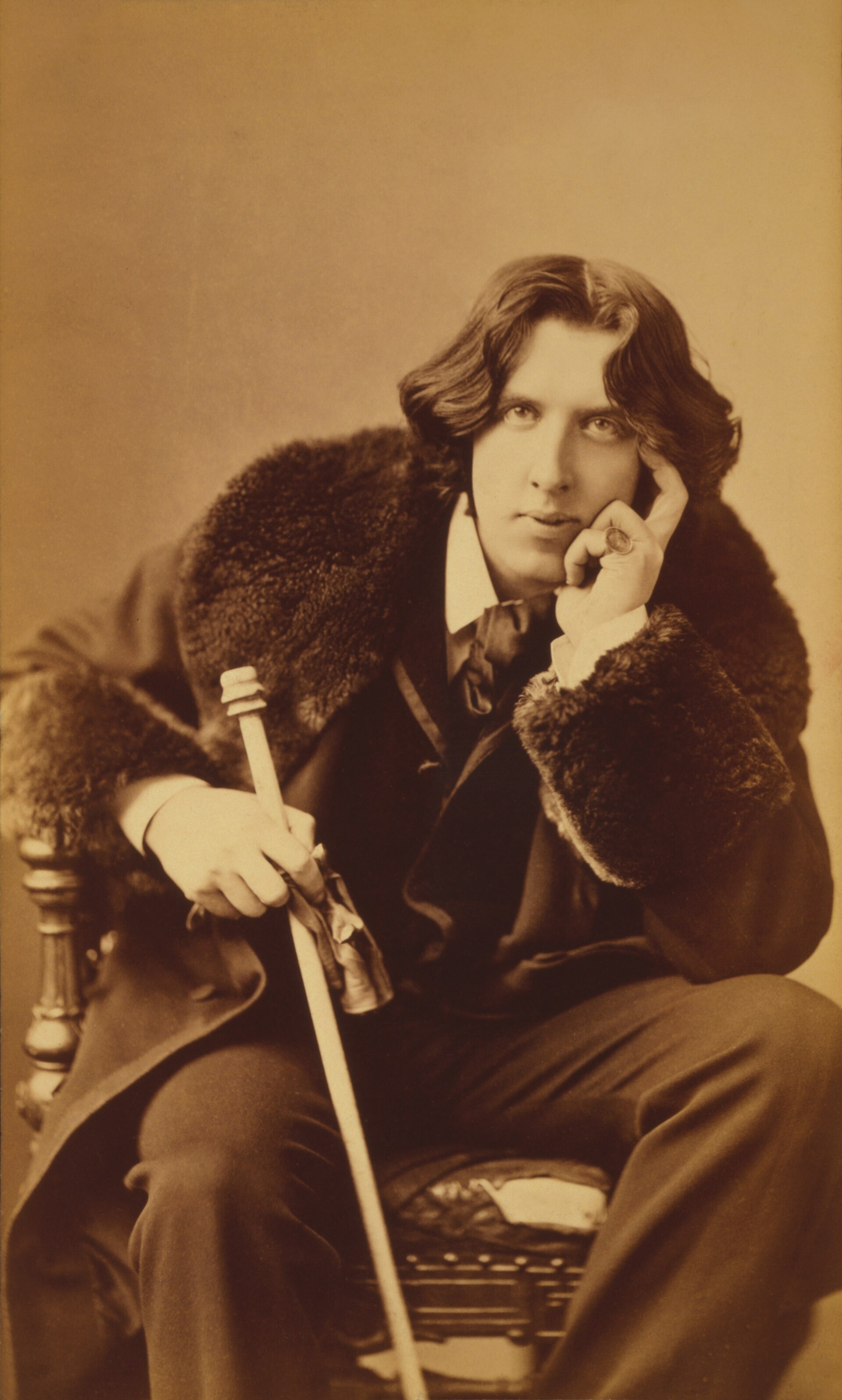 Retrato fotográfico de estudio de Oscar Wilde sentado, con abrigo de piel y bastón, hacia 1882