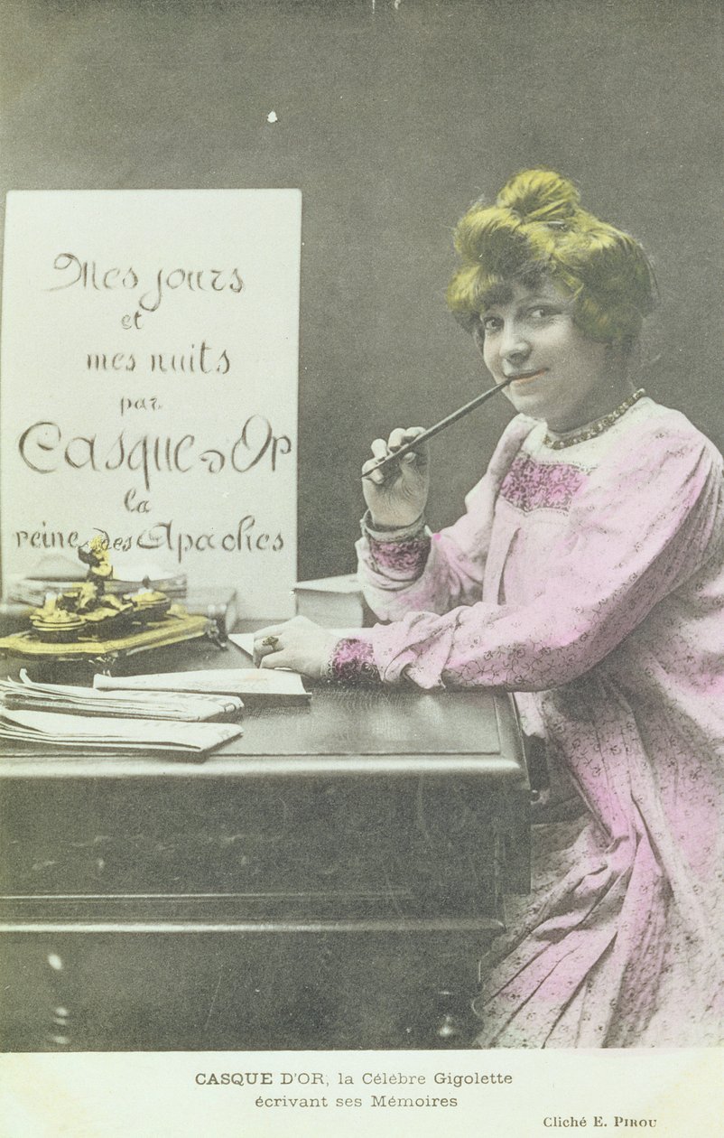 Retrato fotográfico coloreado de hacia 1902 de una mujer joven escribiendo en una mesa, con un cartel que anuncia «Mis días y mis noches, por Casque d'Or, la reina de los apaches»