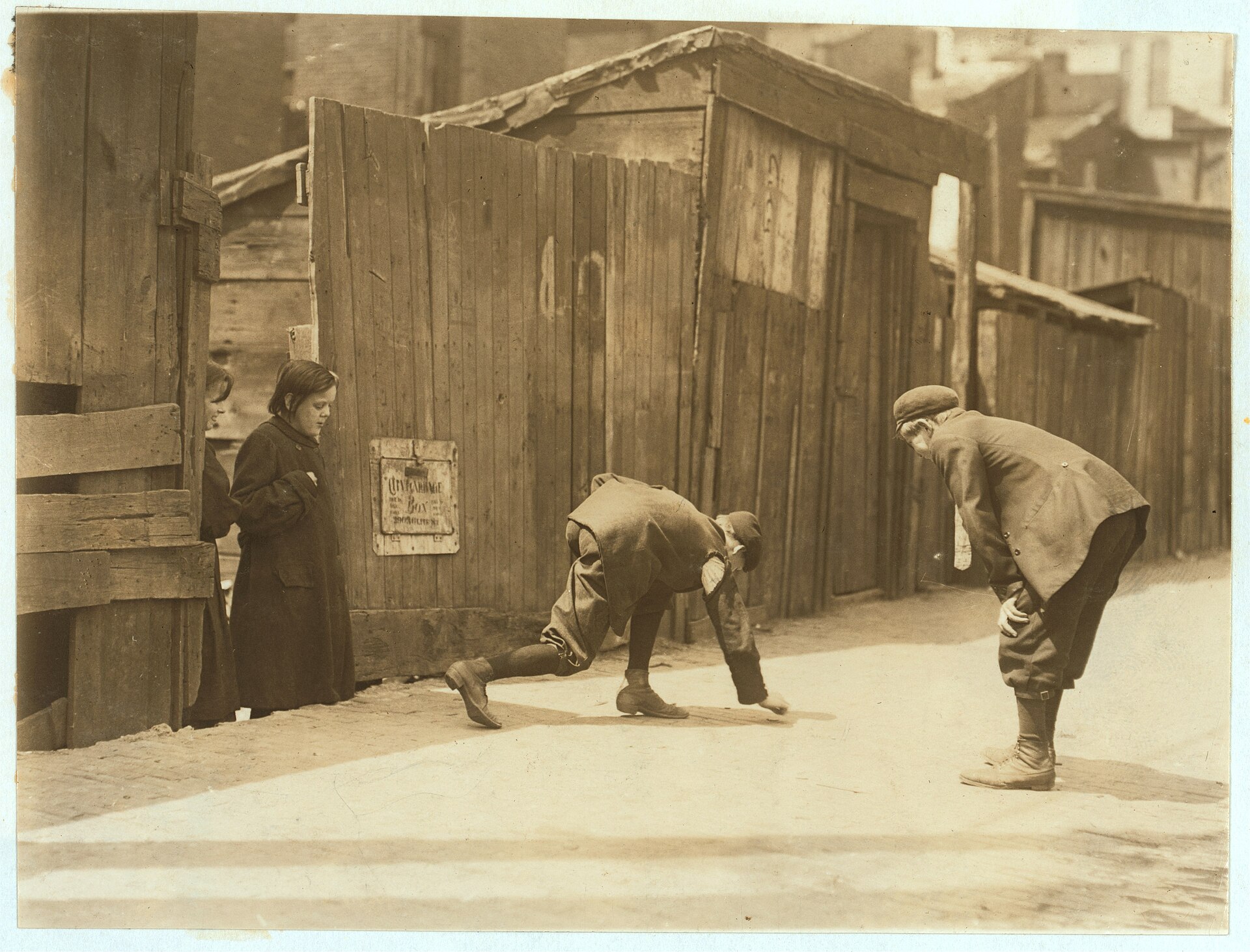 Fotografía de 1910 de dos chicos jugando a los dados en la acera junto a una valla de madera mientras una mujer los observa desde el fondo