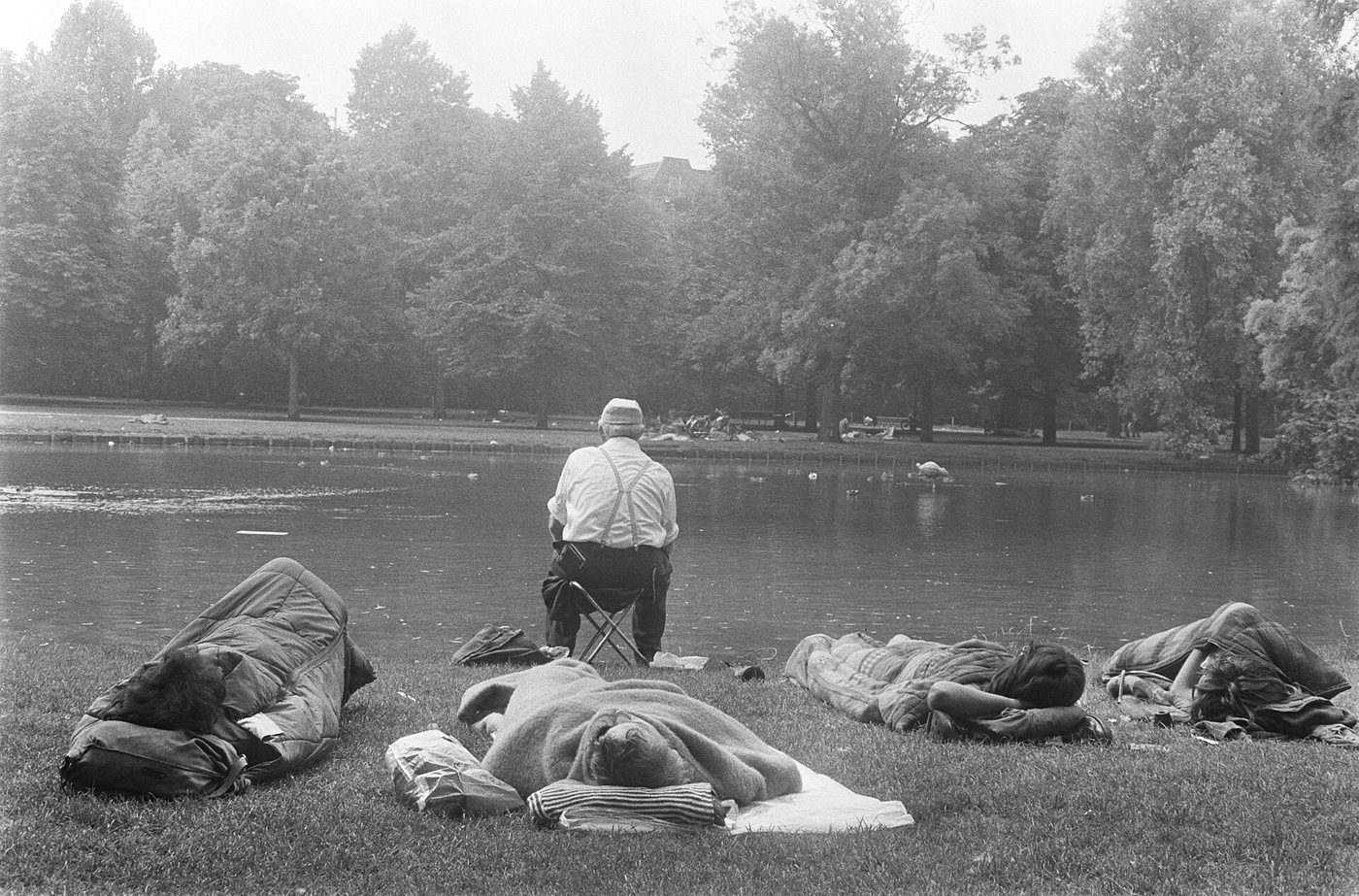 Fotografía de finales de los años sesenta de un grupo de jóvenes durmiendo tumbados en la hierba de un parque mientras un hombre mayor pesca a pocos metros