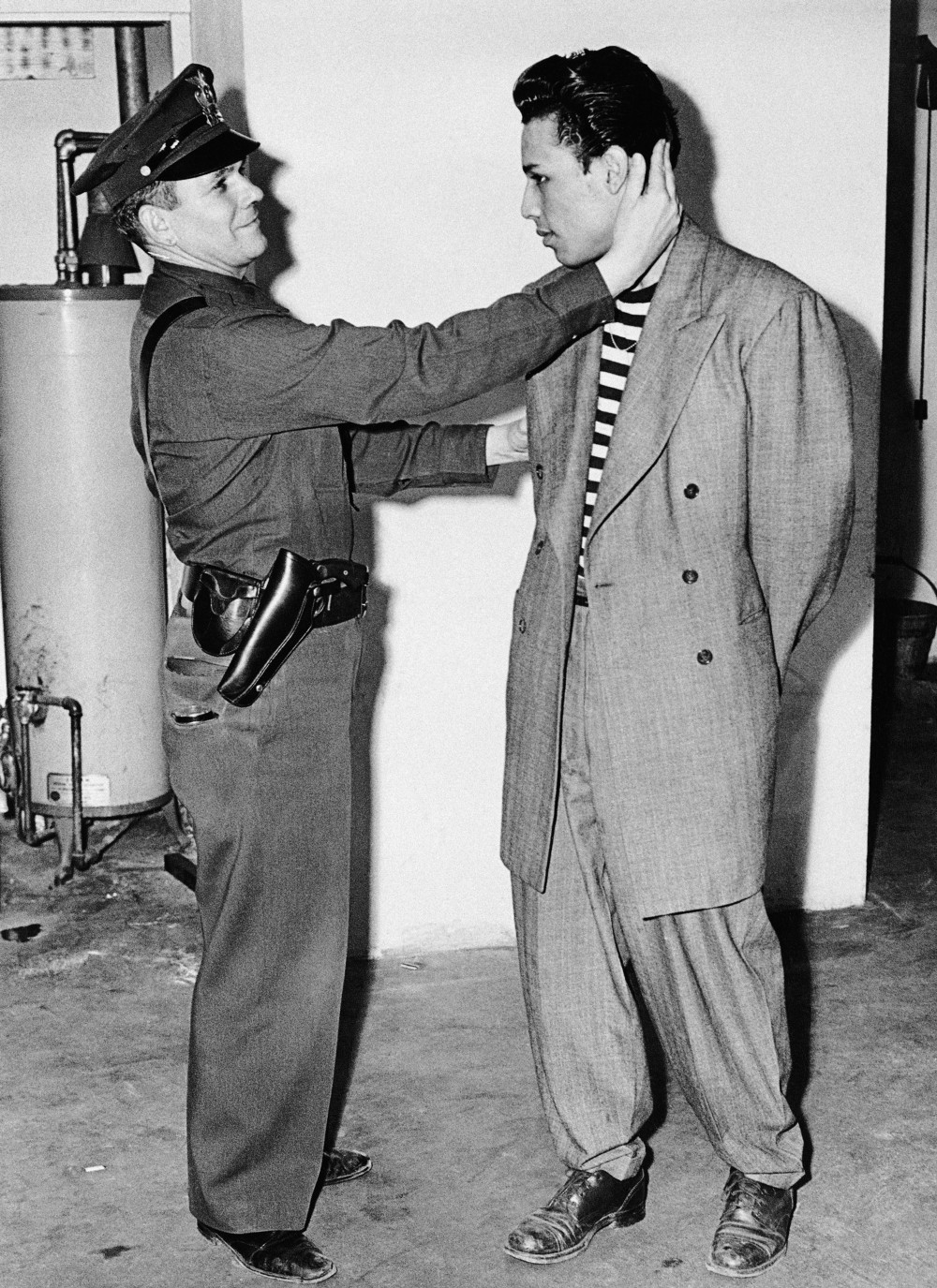 Fotografía de 1943 de un policía inspeccionando la ropa de un joven con traje de pantalón muy ancho y chaqueta larga, contra una pared